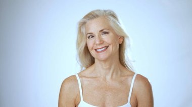 Uzun açık renk saçlı olgun bir kadın spa ortamının keyfini çıkarırken mutlu bir şekilde gülümsüyor. Temiz beyaz bir arka plana karşı rahatlık ve memnuniyet yayıyor..