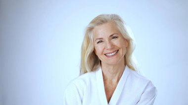 Uzun, açık renk saçlı olgun bir kadın rahatlatıcı spa ortamında eğlenirken mutlu bir şekilde gülümser. Parlak arka plan onun neşeli ifadesini arttırır..