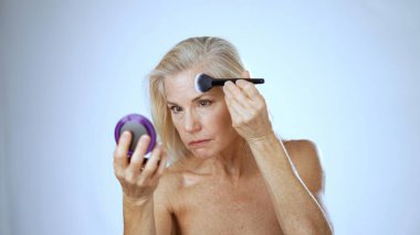 Mutlu, olgun bir kadın makyajını fırçayla yaparken doğal güzelliğini vurgulayarak gülümsüyor. Huzurlu ve aydınlık bir ortamda spa deneyiminin tadını çıkarıyor..