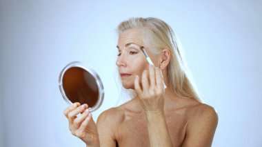 Olgun bir kadın, sakin bir spa ortamında makyaj yaparken mutlu bir şekilde gülümser. Yumuşak arka plan onun neşeli ifadesini vurguluyor ve güzelliğe odaklanıyor..