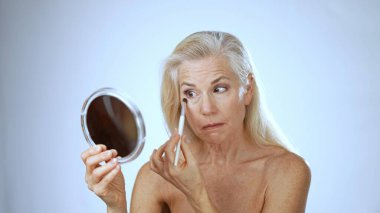 Olgun bir kadın huzurlu bir kaplıcada oturur ve aynaya mutlu bir şekilde makyaj yapar. Rahatlamış tavrı, bu huzurlu ortamda neşe ve özgüveni yansıtıyor..