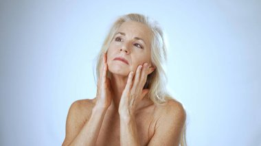 Olgun bir kadın gülümsüyor ve nazikçe yüzüne dokunuyor. Parlak ve rahatlatıcı bir ortamda spa tedavisinin mutluluğunu yaşıyor..