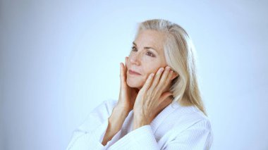 Mutlu, olgun bir kadın, kaplıcada otururken makyaj yaparken gülümser. Parlak arka plan, kendi bakımının barışçıl atmosferine katkı sağlıyor..