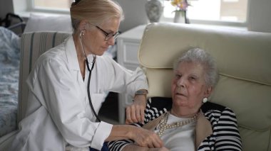 Bakıcı, evde yaşlı bir kadını steteskop kullanarak muayene eder ve rahatını sağlar. Atmosfer sakin, kişiselleştirilmiş sağlık hizmeti sağlıyor..
