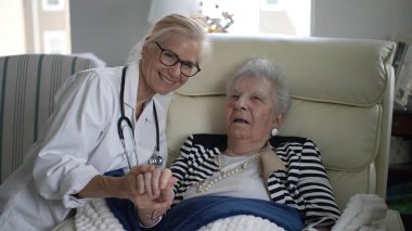 Bakıcı ev ziyareti sırasında yaşlı bir kadınla iletişim kurar, rahat bir oturma odasında rahatlık ve arkadaşlık teklif eder..