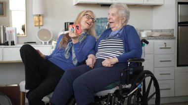 Tekerlekli sandalyedeki yaşlı bir kadın kızıyla mutlu bir şekilde rahat evlerinde akıllı bir telefon kullanarak anılarını ve birlikteliklerini kutluyor..
