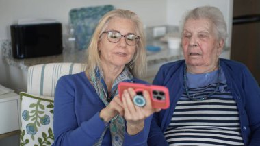 Bir kız, evde rahat bir ortamda yaşlı annesine fotoğraf göstermek için akıllı telefonunu kullanır. Birlikte anıları paylaşıyorlar, sevgiyi ve özeni vurguluyorlar..