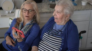 Tekerlekli sandalyedeki yaşlı bir kadın, evine yaptığı samimi bir ziyaret sırasında kızıyla anılarını paylaşırken sevgi ve desteğin özünü yakalıyor..