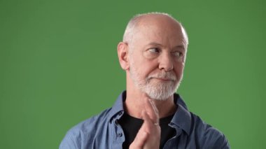 Sakallı orta yaşlı bir adam mavi bir gömlek giyer ve yeşil perde arkasında dururken düşünceli hareketler sergiler. Bir anlık tefekkür kaydedildi..
