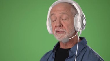 Sakallı orta yaşlı bir adam yeşil ekran arka planında bir kulaklık ve mikrofon takıyor. Özenli ve meşgul görünüyor, muhtemelen sohbet halinde..