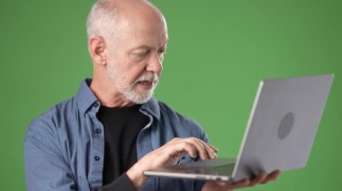 Orta yaşlı bir adam dizüstü bilgisayardaki içeriği açıklarken jest yapar, yeşil bir perdeye dayanır, ifadesindeki bağlılık ve berraklığı gösterir..
