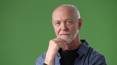 Sakallı olgun bir adam yeşil bir ekrana karşı duruyor. İzleyiciyi dostça bir ifadeyle işaret ediyor. Bağlılık ve bağlılık duygusunu iletiyor..