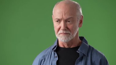 Olgun bir adam yeşil bir ekranın önünde duruyor, eli boğazına yakın bir şekilde endişeyi açıkça ifade ediyor, ciddi bir duygu ya da mesaj iletiyor..