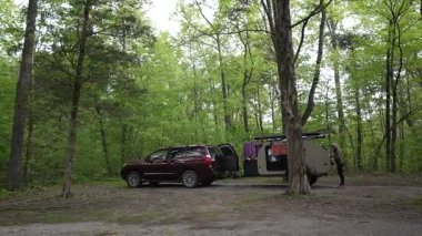 Bir kadın ailesiyle birlikte manzaralı bir ormanda yaz kampı gezisinden zevk alır. Yakınlarda yeşil ağaçlarla çevrili bir gözyaşı römorku var..