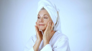 Olgun bir kadın, huzurlu bir spa ortamında mutlu bir şekilde gülümserken beyaz bir bornoz ve havlu giyer, gençleştirici deneyimi sırasında rahatlama ve neşe saçar..