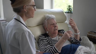 Yaşlı bir kadın evde bakıcısıyla sohbet eder, bir sandalyede rahat bir şekilde otururken düşüncelerini paylaşır ve rahat atmosferin tadını çıkarır..