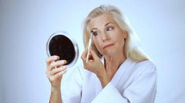 Kaplıcada küçük bir ayna kullanarak makyaj yaparken gülümseyen olgun bir kadın. Arka plan parlak ve sakin, rahatlamasını sağlıyor..