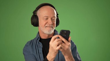 Son sınıf öğrencisi, akıllı telefonuyla büyük kulaklıklar takarak mutlu bir şekilde iletişim kurar. Katı yeşil arka plan onun neşeli ifadesini vurguluyor ve müziğe odaklanıyor..