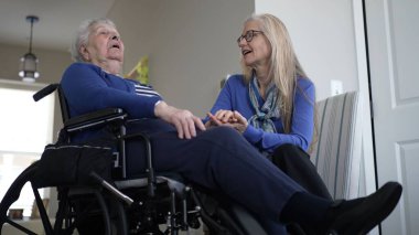 Tekerlekli sandalyedeki yaşlı bir kadın kızıyla sıcak bir ziyaretten hoşlanıyor. Sıcak ev ortamlarında sevgi ve bakım anlarını paylaşıyorlar..