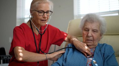 Konforlu bir sağlık merkezinde, bakıcı yaşlı bir kadına bir anlık bağlantı ve bakımı paylaşırken özenle yardımcı olur. Atmosfer bağlılık ve rahatlığı teşvik eder..