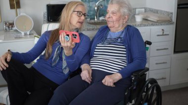Tekerlekli sandalyedeki yaşlı bir kadın kızıyla görüntülü sohbetten hoşlanıyor. İkisi de rahat bir evde anılarını paylaşırken gülümsüyor..