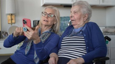 90 'lı yaşlarda bir kadın tekerlekli sandalyede otururken, kızı ona akıllı telefondan bir şey gösteriyor, evde sevgi ve destek anı yakalıyor..