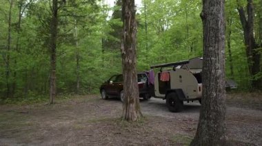 Bir kadın ailesiyle birlikte manzaralı bir ormanda yaz kampı gezisinden zevk alır. Yakınlarda yeşil ağaçlarla çevrili bir gözyaşı römorku var..