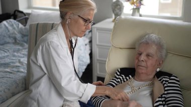 Olgun bir kadın rahat evindeki bir hemşireden sağlık kontrolü alır. Hemşire steteskop kullanırken kadın rahat bir şekilde koltukta oturuyor..