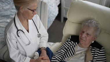 Beyaz önlüklü bir hemşire, evde rahat bir sandalyede oturan yaşlı bir kadına arkadaşlık ve bakım sunar..