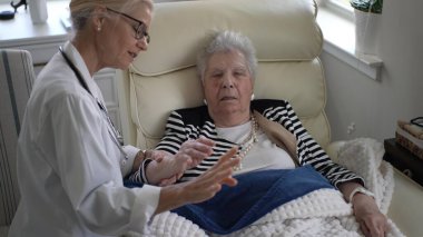 Olgun bir kadın rahat bir sandalyede dinlenirken bir bakıcı dikkatlice sağlığını tartışıyor. Ortam sıcak ve şefkatli, ziyaret sırasında rahatlık sağlıyor..
