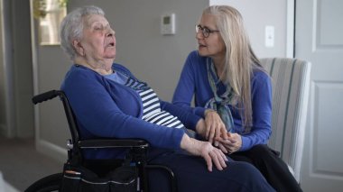 Bir kız çocuğu, eve yaptığı özel bir ziyaret sırasında yaşlı annesine yakın bir tekerlekli sandalyede sevgi ve destek sunuyor. Hava sıcak ve şefkatli..