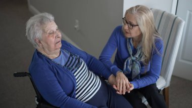 Tekerlekli sandalyedeki yaşlı bir kadın ziyaret sırasında kızından sevgi dolu destek alır. Sıcak, kapalı bir mekanda birlikte hassas bir anı paylaşıyorlar..