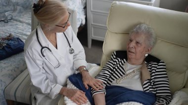 Yetişkin bir kadın evdeki bir sandalyede rahatça otururken, bir sağlık uzmanı onunla konuşarak ziyaret sırasında arkadaşlık ve tıbbi tavsiyelerde bulunuyor..