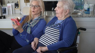 90 'lı yaşlarda bir kadın tekerlekli sandalyede otururken, kızı ona akıllı telefondan bir şey gösteriyor, ev ortamında neşeli anılarını paylaşıyor..