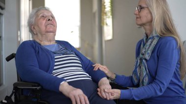 90 'lı yaşlarda yaşlı bir kadın, günlük ziyareti sırasında kızından sevgi dolu bir destek alarak evinde tekerlekli sandalyede oturuyor. İçerideki atmosfer sıcak ve davetkar..