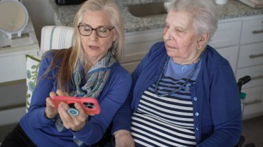 60 'lı yaşlarda bir kadın, 90' lı yaşlardaki yaşlı annesine sıcak bir mutfakta oturup, anılarını paylaşırken ve birlikte vakit geçirirken akıllı telefon kullanmasında yardımcı olur..