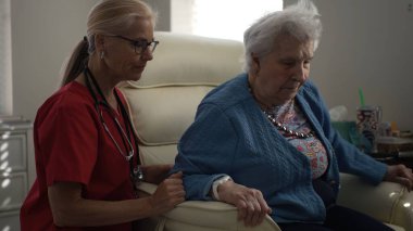 Önlüklü bir bakıcı, bir sandalyede oturan yaşlı bir kadınla iletişim kurar, ev gibi bir sağlık ortamında ilişki ve duygusal destek sağlar..