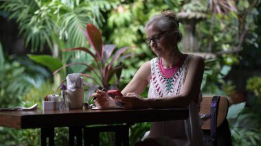 Orta yaşlı bir kadın, canlı tropikal bitkilerin ortasında huzurlu bir anın tadını çıkarırken bir kafe masasında cihazına odaklanmış şekilde oturuyor..