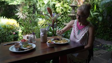 Orta yaşlı bir kadın gür yeşilliklerle çevrili canlı bir kafede oturuyor. Bilgisayarı üzerinde çalışırken, dijital göçebe yaşam tarzını somutlaştırarak lezzetli bir yemek yiyor..