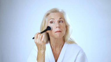 Cübbe giymiş mutlu, olgun bir kadın, hafif arka planlı, sakin bir kaplıca ortamında otururken ruj sürerken gülümsüyor..