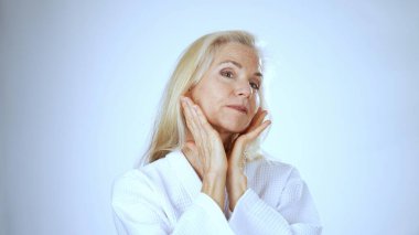 Beyaz cüppeli mutlu olgun bir kadın, sakin ve parlak bir arka planla çevrili aynaya bakarken makyaj yapar. Neşe ve güven saçıyor.