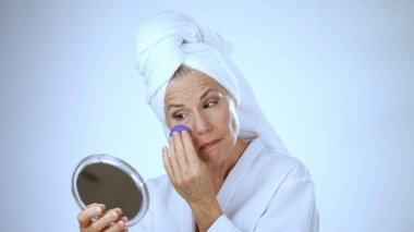 Kafasında havlu olan olgun bir kadın parlak bir spa ortamında cilt bakımı yaparken hoş bir şekilde gülümsüyor. Onun mutlu ifadesi rahatlamayı ve eğlenmeyi öneriyor..