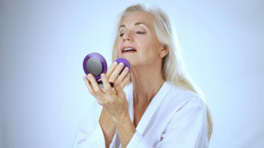 Kaplıca ortamında mutlu, olgun bir kadın aynadaki yansımasına bakar. Huzurlu, beyaz bir arka planda neşe ve rahatlama yaşar..