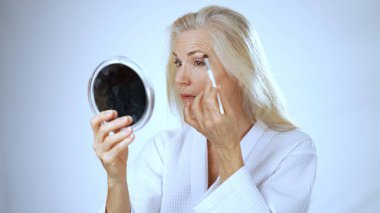 Olgun bir kadın spa ortamında mutlu bir şekilde gülümser, fırçayla makyaj yapar. Huzurlu beyaz arka plan onun neşeli ifadesini ve kişisel bakımını arttırır..