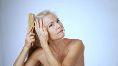 Mutlu olgun bir kadın uzun saçlarını nazikçe tararken kameraya gülümser. Sakin, beyaz bir arka planı olan, huzur saçan bir kaplıca ortamında..