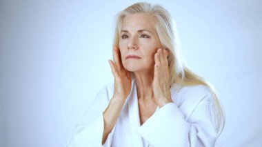 Olgun bir kadın, huzurlu bir kaplıca arka planına cilt bakımı yaparken sevinçle gülümser. Mutluluğu beyaz bir bornozla yayıyor ve ferahlatıcı atmosferi kucaklıyor..