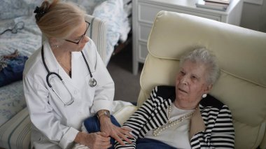 Bir sağlık hizmetleri uzmanı, yaşlı bir kadına evinde bakım sağlıyor. Oturma odasında rahatça otururken sohbet ediyorlar..