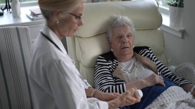 Olgun bir kadın, bir sağlık hizmeti sağlayıcısı ev ziyareti sırasında ilaç detaylarını tartışırken rahat bir şekilde sandalyeye oturur. Şefkat ve şefkat aşikar..