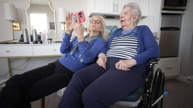 Bir kız, evde rahat bir şekilde otururken akıllı telefon kullanarak, birbirleriyle olan bağlarını ve ilgilerini göstererek, yaşlı annesiyle neşeli anlar paylaşır..