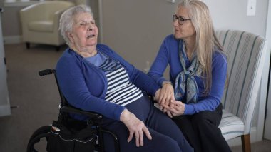 Kızı sevgi dolu bir şekilde tekerlekli sandalyedeki yaşlı annesiyle el ele tutuşuyor, samimi bir ziyaret sırasında ona bakıcılık ve destek yapıyor, yakın bağlarını gösteriyor..